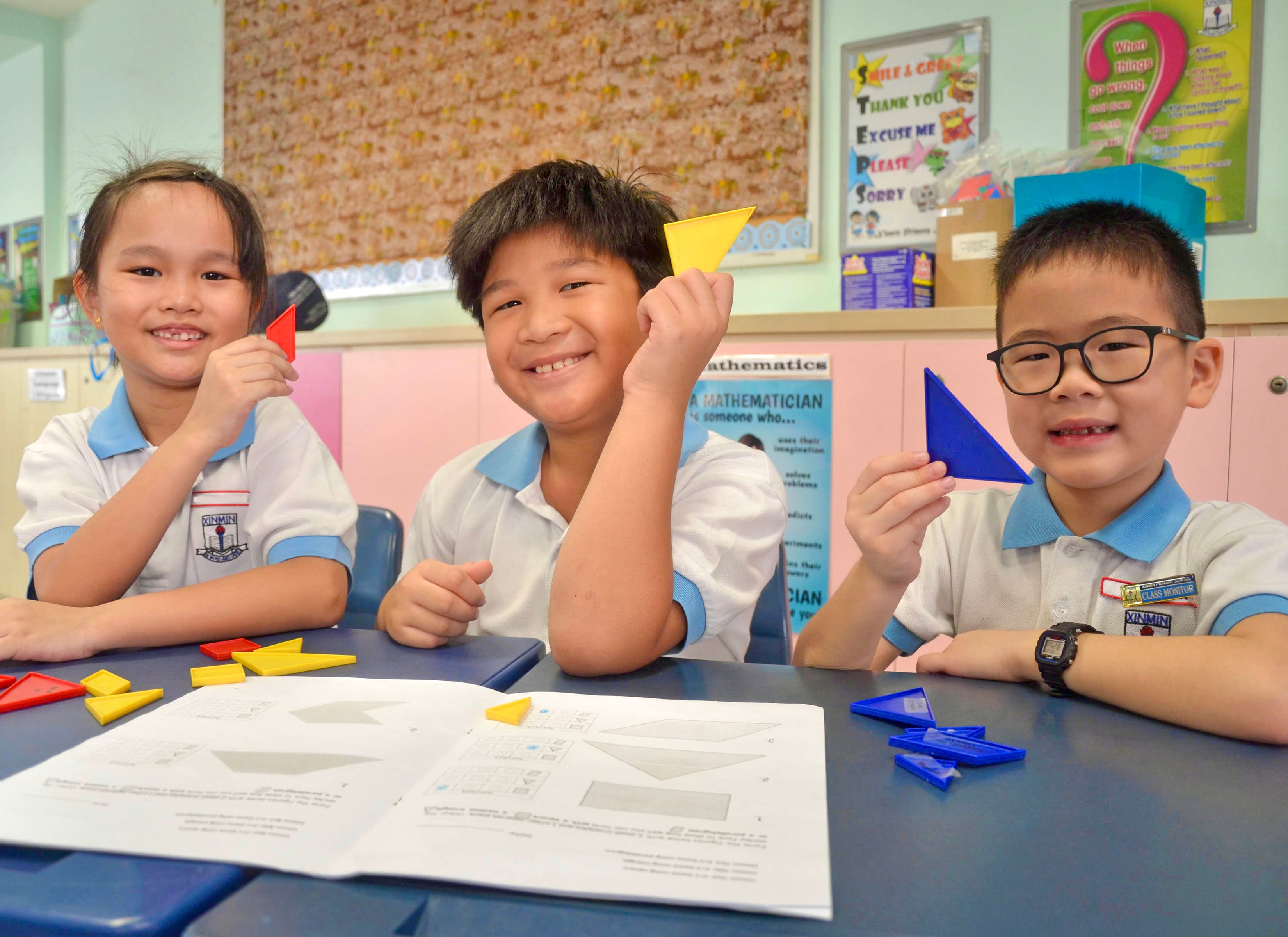 Three smiling children in school uniforms hold colorful plastic triangles at a classroom desk with worksheets.